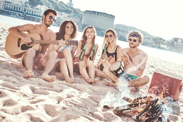 Grupo de amigas jóvenes sentados en la playa en hamacas cantando y tocando la guitarra