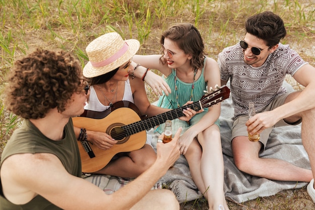 Grupo de amigos alegres pasar tiempo juntos al aire libre, bebiendo cervezas, tocando la guitarra