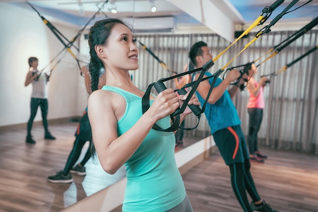 grupo de amigos asiáticos haciendo ejercicios de resistencia corporal total. los deportistas entrenan en interiores con trx. mujeres musculosas y hombres haciendo ejercicio en el aula de gimnasia con espejos alrededor.