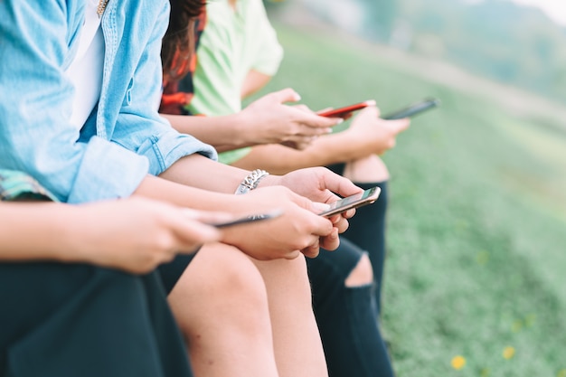 Un grupo de amigos asiáticos recortados están sentados usando sus teléfonos inteligentes y charlando en el parque