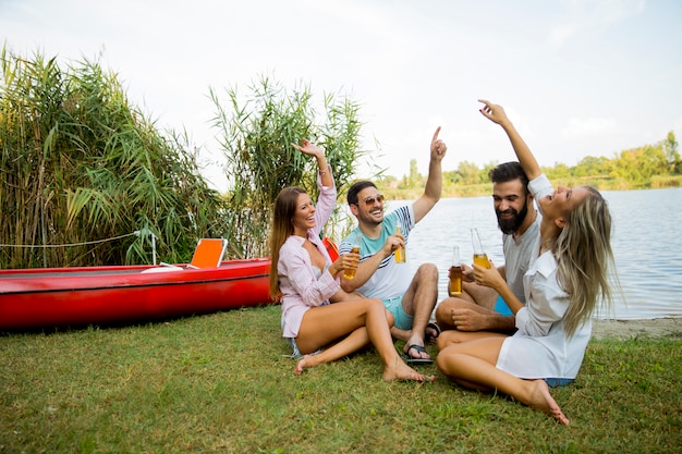 Grupo de amigos con botellas de sidra sentado junto al barco cerca del hermoso lago y divirtiéndose