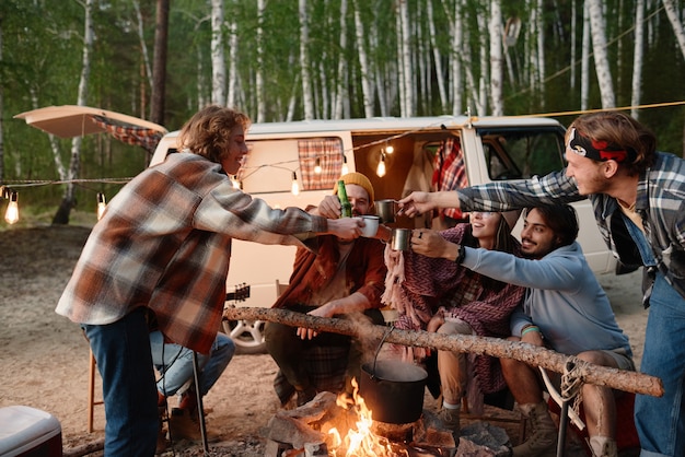 Grupo de amigos brindando junto con bebidas cerca del fuego durante el campamento en el bosque