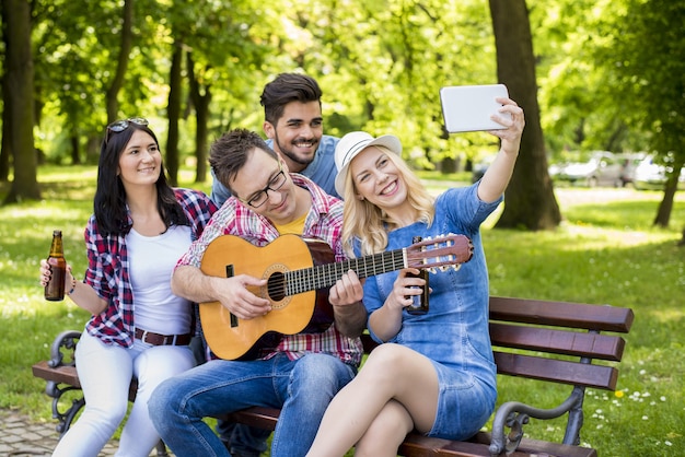 Grupo de amigos caucásicos tocando la guitarra y tomando selfies en un banco del parque