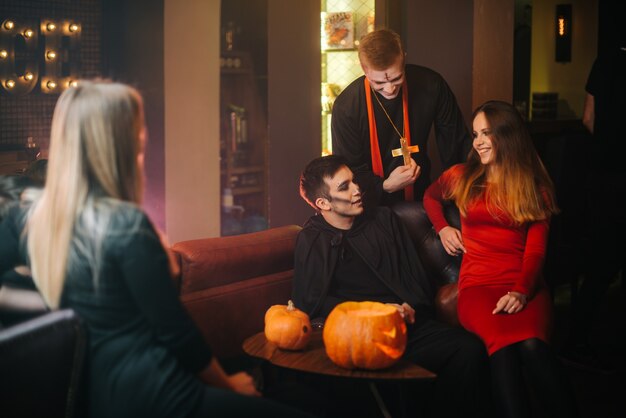 Foto grupo de amigos celebran halloween en la cafetería. guy está vestido como un terrible sacerdote y monstruo. hermosa chica sexy con un vestido rojo sentado en el sofá. disparos en una habitación con paredes negras y rojas.