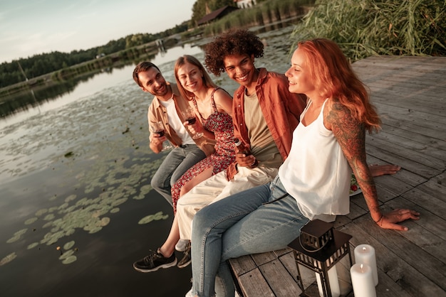 Grupo de amigos divirtiéndose en un picnic cerca de un lago, sentados en el muelle de madera comiendo y bebiendo vino, cerveza, sidra. Jóvenes sonrientes con celebración de fiestas al aire libre durante la puesta de sol en el campo