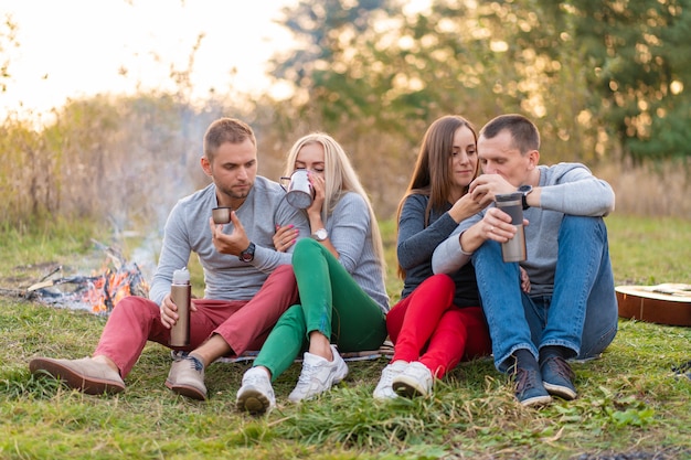 Un grupo de amigos está disfrutando de una bebida caliente de un termo, en una tarde fresca junto a un incendio en el bosque. Tiempo divertido para acampar con amigos