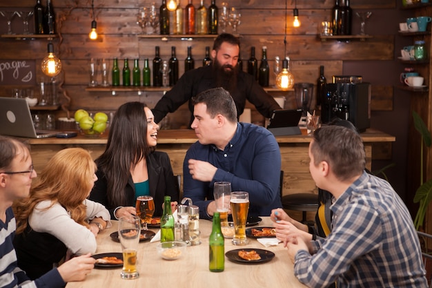 Grupo de amigos felices bebiendo cerveza y comiendo pizza en el bar restaurante. Están sentados en una mesa de madera.