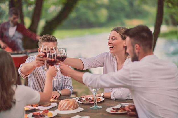 grupo de amigos felices brindando con una copa de vino tinto mientras hacen un picnic en una cena francesa al aire libre durante las vacaciones de verano cerca del río en la hermosa naturaleza