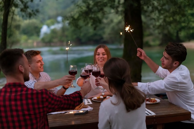 grupo de amigos felices brindando con una copa de vino tinto mientras hacen un picnic en una cena francesa al aire libre durante las vacaciones de verano cerca del río en la hermosa naturaleza