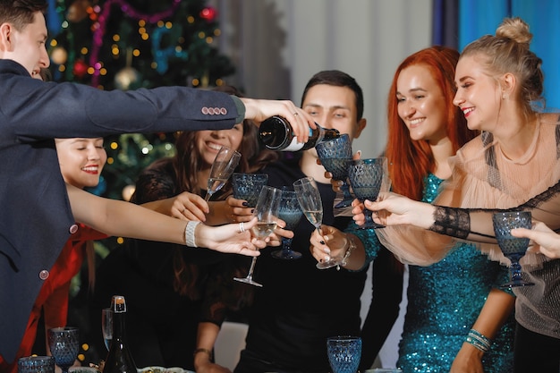 Grupo de amigos felices celebrando la Navidad o el año nuevo. Un hombre vierte champán en copas. Las mujeres ríen, sentadas en una mesa cerca del árbol de Navidad.