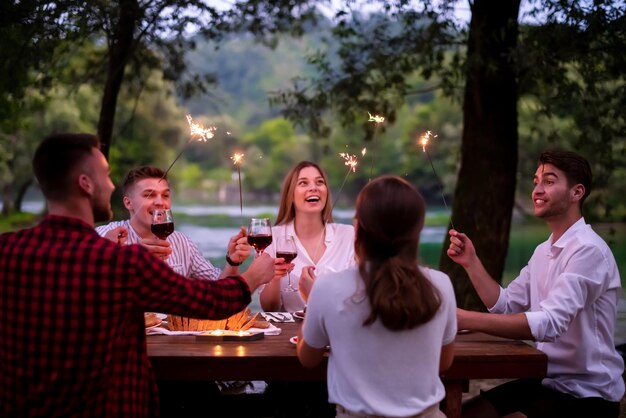 grupo de amigos felices celebrando las vacaciones usando rociadores y bebiendo vino tinto mientras cenan en un picnic francés al aire libre cerca del río en una hermosa tarde de verano en la naturaleza
