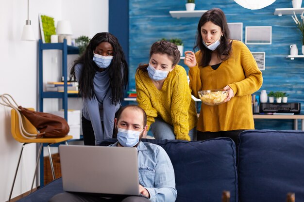 Grupo de amigos multiétnicos que mantienen el distanciamiento social con una mascarilla para evitar la propagación del coronavirus mirando una computadora portátil en la sala de estar de su casa y pasan tiempo juntos bebiendo cerveza comiendo papas fritas. Conc