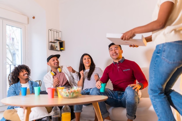 Grupo de amigos multiétnicos en un sofá comiendo pizza y bebiendo refrescos en una fiesta en casa muy felices recibiendo a la chica con la pizza