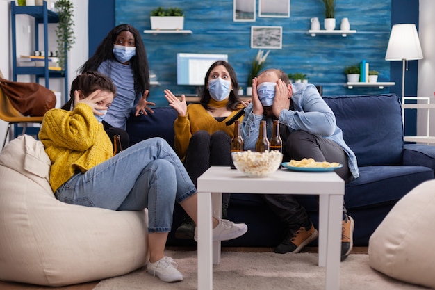 Grupo de amigos multiétnicos viendo una película de terror en la televisión teniendo miedo de usar mascarilla para prevenir la infección con covid 19 durante la pandemia global divirtiéndose sentado en el sofá y