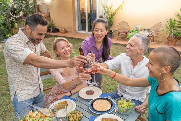 Grupo de amigos multirraciales brindando con una hermosa sonrisa alrededor de la mesa en el comedor del patio de la casa