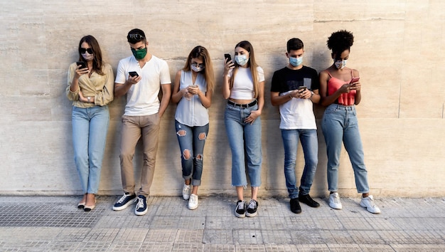 Grupo de amigos multirraciales con máscaras faciales frente a una pared limpia usando teléfonos inteligentes para compartir contenido en las redes sociales. Covid-19 y concepto de estilo de vida tecnológico