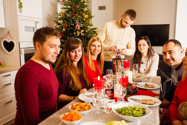 Grupo de amigos sentados en la mesa en la víspera de Navidad. Selebrando juntos Fiesta de Año Nuevo, comidas y bebidas. Champán abierto hombre