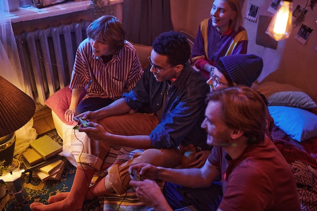 Grupo de amigos sentados en el suelo en una habitación oscura con joysticks mientras juegan videojuegos en una fiesta