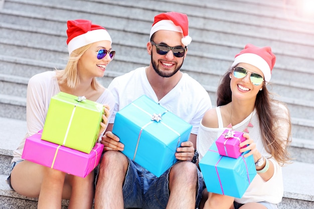 Grupo de amigos con sombreros de Papá Noel sentados en la ciudad con regalos