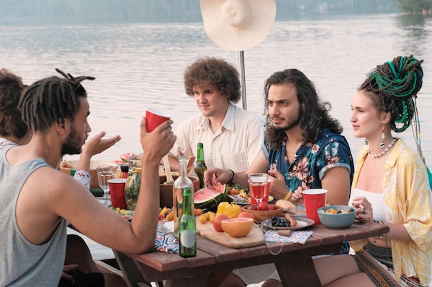 Grupo de amigos tienen un picnic en la naturaleza que están sentados en la mesa de comedor comiendo, bebiendo y hablando