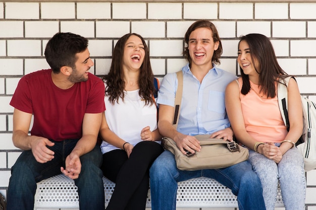 Grupo de amigos universitarios latinos hablando y divirtiéndose mientras están sentados en un banco en el pasillo de la escuela