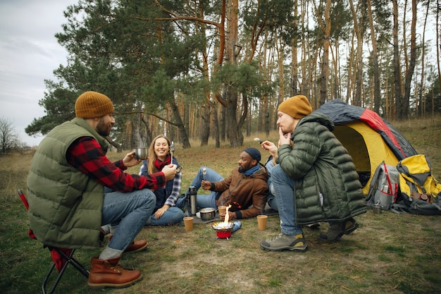 Grupo de amigos en un viaje de campamento o senderismo en día de otoño. Hombres y mujeres con mochilas turísticas tomando un descanso en el bosque, hablando, riendo. Actividad de ocio, amistad, fin de semana. Comer y beber.