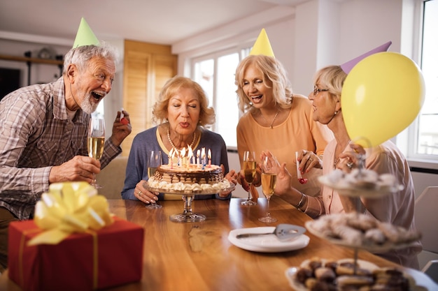 Grupo de ancianos alegres divirtiéndose en la fiesta de cumpleaños mientras la mujer sopla velas en un pastel