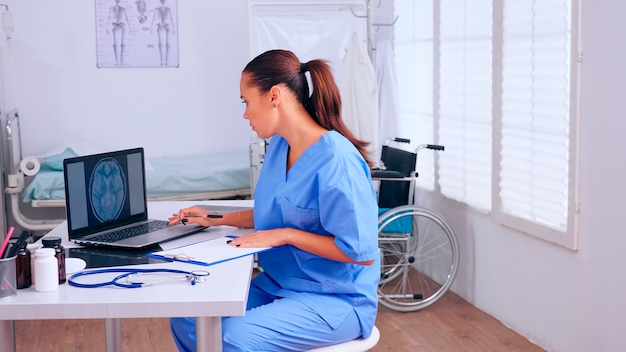 Grupo de asistentes que trabajan en la oficina del hospital, analizando radiografía cerebral digital y escaneo corporal. Médico en medicina uniforme escribiendo una lista de pacientes consultados y diagnosticados, realizando una investigación.