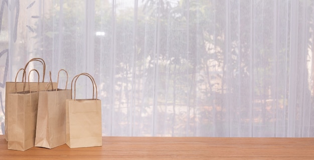 Grupo de bolsas de papel kraft con espacio de copia en mesa de madera con ventanas de cortina blanca.