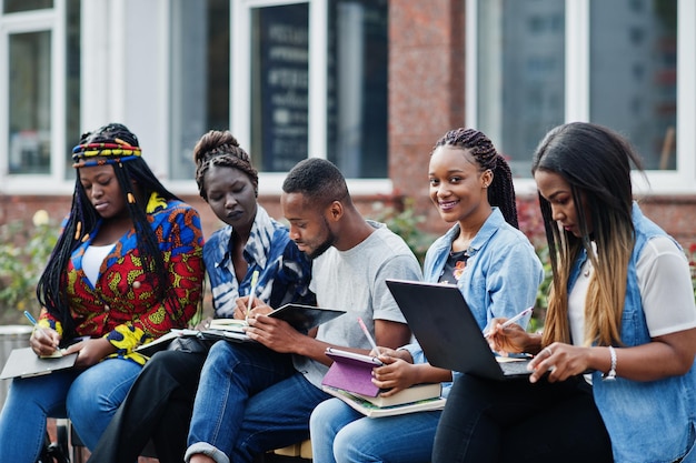 Grupo de cinco estudiantes universitarios africanos que pasan tiempo juntos en el campus en el patio de la universidad Amigos afro negros que estudian en un banco con artículos escolares computadoras portátiles cuadernos