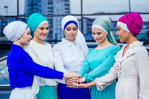 Un grupo de cinco jóvenes y atractivas mujeres de negocios con vestido musulmán hijab multicolor y un turbante que cubre la cabeza, trabajando con los pulgares hacia arriba, apilando las manos, junta la palma de la mano en el trabajo en equipo del centro.