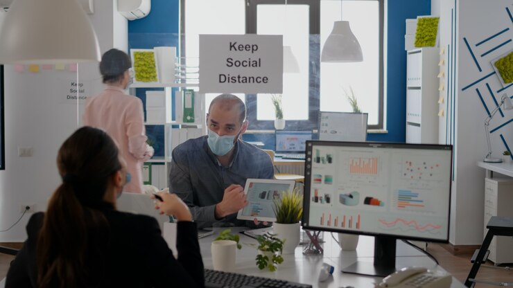 Foto grupo de compañeros de trabajo de negocios con máscaras analizando gráficos con tableta digital sentado en el nuevo lugar de trabajo normal. colega que trabaja en segundo plano mantiene el distanciamiento social