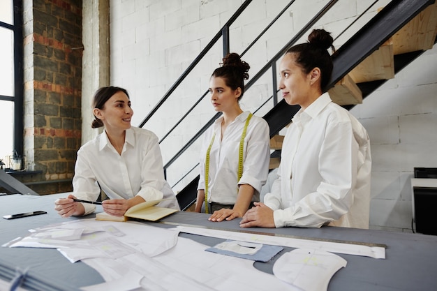 Grupo de creativos diseñadores de moda modernos en camisas blancas intercambiando ideas y discutiendo ideas para la nueva colección de temporada en la reunión