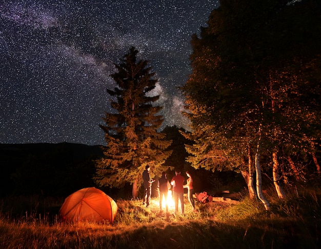 Grupo de cuatro amigos excursionistas calentándose las manos alrededor de una fogata, descansando cerca de una carpa naranja brillante bajo un cielo inusualmente estrellado