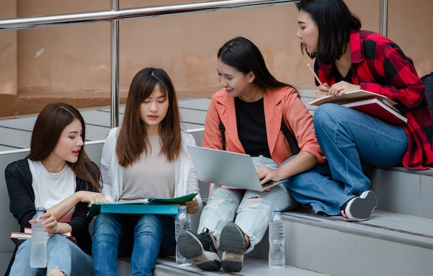 Grupo de cuatro atractivos estudiantes universitarios asiáticos sentados en cada escalera en el campus universitario que estudian al aire libre. Concepto de educación, amistad y vida de estudiantes universitarios.