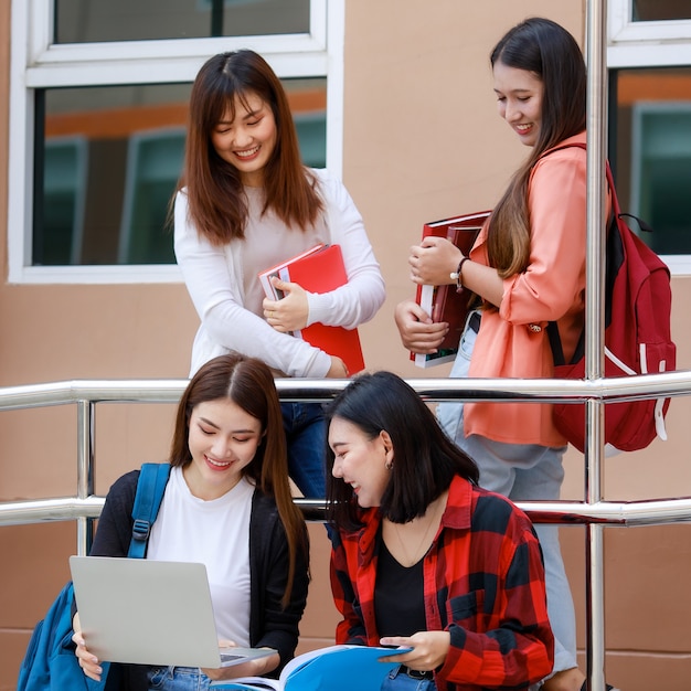 Grupo de cuatro chicas estudiantes universitarias con libros y ordenador portátil sentado y hablando.