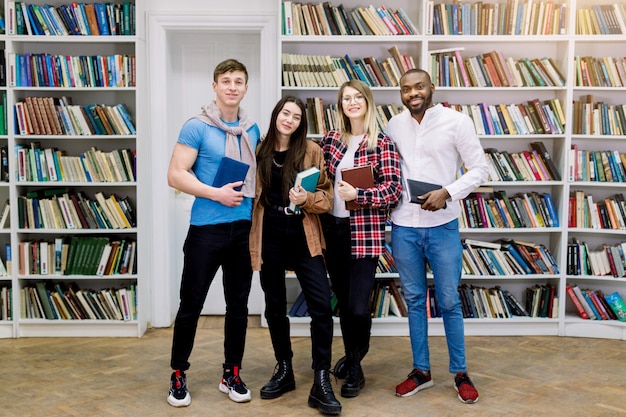 Grupo de cuatro estudiantes multiétnicos sonrientes confiados, niñas y niños en ropa casual, posando en la biblioteca, sosteniendo libros y mirando a camer