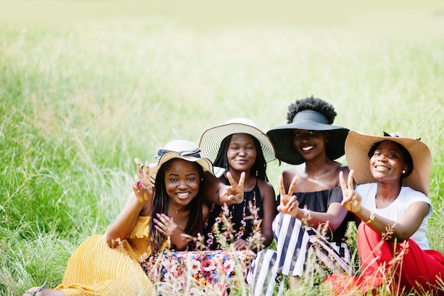 Grupo de cuatro hermosas mujeres afroamericanas usan sombrero de verano sentado en la hierba verde en el parque.