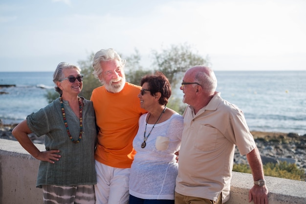 Grupo de cuatro personas maduras divirtiéndose y hablando juntos en la playa - jubilados ancianos sonriendo y riendo con el mar al fondo