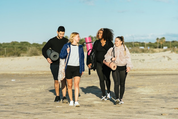 Grupo de cuatro personas multiétnicas caminando por la playa con colchonetas y bolsas de yoga