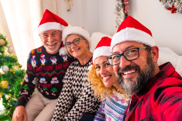 Grupo de cuatro personas sonriendo y mirando a la cámara del joven sosteniendo la cámara - selfie familiar en casa el día de Navidad juntos