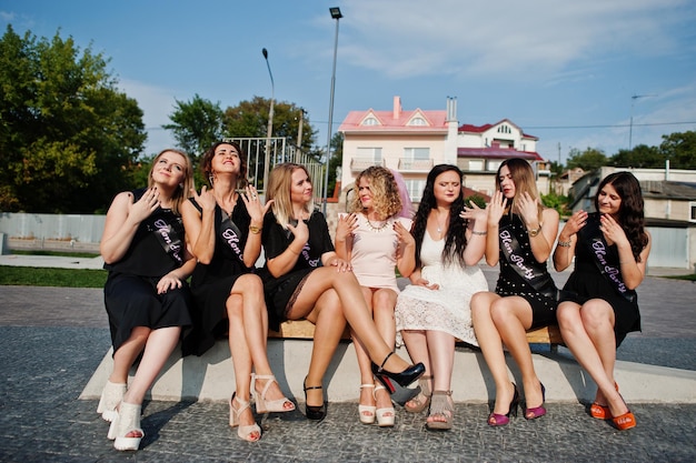 Grupo de 7 meninas vestem preto e 2 noivas sentadas no banco e se divertindo na festa de despedida