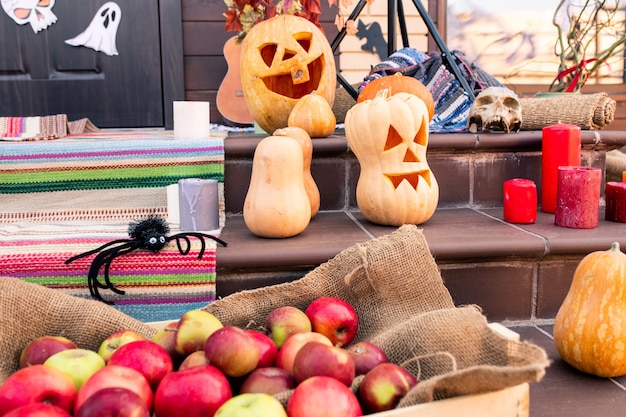 Grupo de abóboras maduras de halloween, aranha, pilha de maçãs, caveira, velas vermelhas na escada e outras coisas pela porta da casa de campo