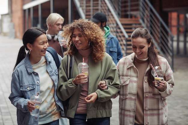 Grupo de adolescentes interculturais alegres com refrigerante conversando ao ar livre