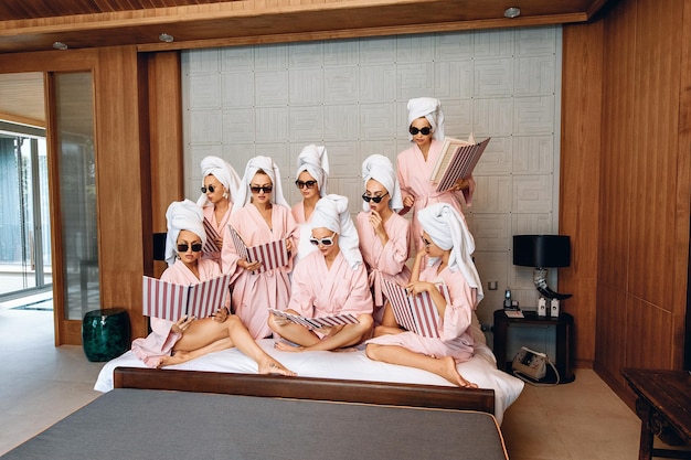 Grupo de adoráveis modelos femininas focadas nas mesmas roupas, vestindo roupões de banho, uma toalha na cabeça e óculos escuros e posando com um menu em suas mãos indoor. Conceito de autocuidado