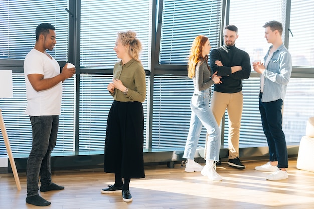 Grupo de alegres colegas multiétnicos, fazendo uma pausa para o café no trabalho, no fundo da janela e a luz solar intensa. equipe de negócios jovem passar algum tempo juntos em um escritório moderno, conversando e discutindo.