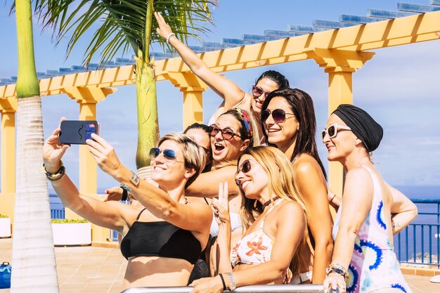 Grupo de amigas aproveitando as férias de verão no resort e tirando selfie usando o smartphone. Mulheres de maiô, aproveitando a festa na piscina. Amigas atraentes tirando fotos com a câmera do celular