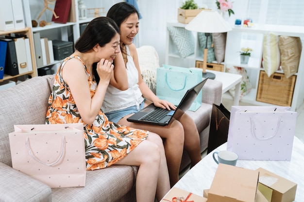 grupo de amigas lindas senhoras sentadas no sofá comprando site de internet on-line no computador portátil rindo sorrindo olhando para a tela. duas mulheres rodeando por caixas de papelão e pacotes de sacos