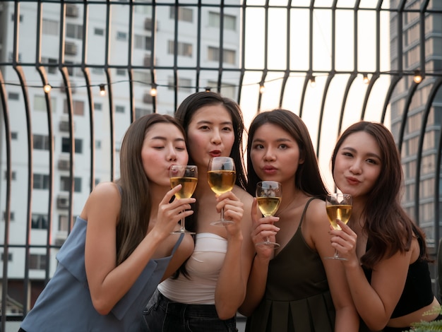 Grupo de amigo asiático torcendo e bebendo na festa no terraço. Jovens brindando copo com vinho no restaurante do último piso