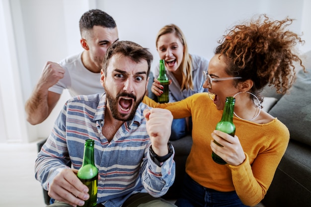 Grupo de amigo sentado na sala e torcendo por seu time de futebol favorito. todos eles segurando garrafas de cerveja.